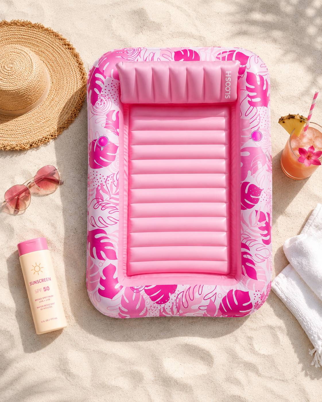 Pink inflatable tanning pool float with floral design on a sandy beach with a sun hat, sunglasses, sunscreen, and drink.